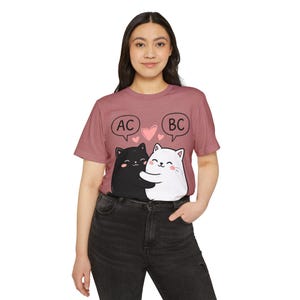 May include: A mauve t-shirt with a graphic of two cartoon cats, one black and one white, embracing. Pink hearts and speech bubbles above the cats read "AC" and "BC". The shirt is made of a soft material.