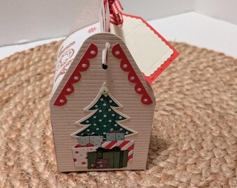 May include: A festive, house-shaped gift box with a Christmas tree and wrapped presents design. The box is beige with red scalloped trim and a red and white checkered ribbon. A blank, red-edged tag is attached.