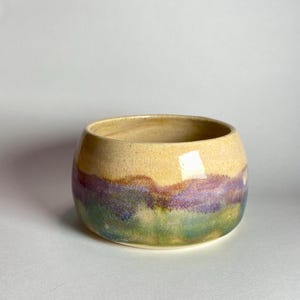 May include: A small, round ceramic bowl with a wide opening. The bowl features a color gradient, with a tan top, transitioning to purple, green, and yellow at the base. The bowl is likely handmade and suitable for various uses.