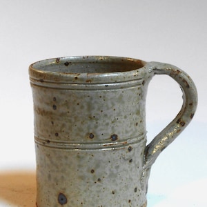 Peut inclure: Une tasse en céramique grise avec un glaçage moucheté et une anse brune. La tasse a un design simple et rustique.