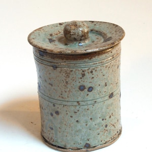 Peut inclure: Un pot en céramique fait à la main avec un couvercle. Le pot est cylindrique avec un glaçage vert clair moucheté et des accents bruns. Le couvercle a une poignée en forme de bouton. Le pot a un aspect rustique et artisanal.