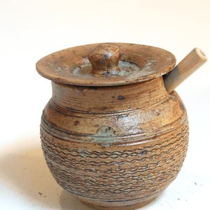 May include: A brown ceramic honey pot with a lid and a wooden dipper. The pot has a textured surface and a brown glaze.