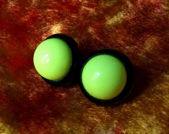 Op de afbeelding: Twee neon groene, bolvormige oordopjes met zwarte randen. De oordopjes staan tegen een getextureerde, roodbruine achtergrond. De oordopjes zijn een helder, opvallend accessoire.