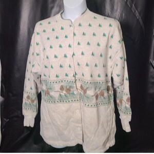 May include: A cream-colored long-sleeve button-down shirt featuring a repeating pattern of small green pine trees on the upper section and a horizontal band across the middle displaying brown pinecones and green pine needles on branches.