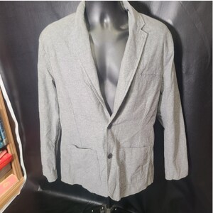 May include: A light gray blazer featuring a notched lapel, two dark buttons for closure, and two large patch pockets on the front. The garment is constructed from a textured fabric and includes a single chest pocket on the left side.