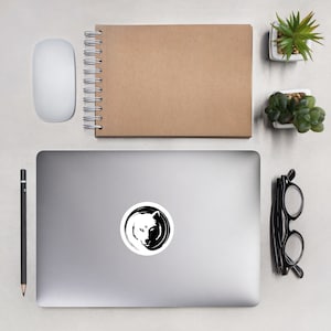 May include: A black and white yin yang symbol with a wolf's head in the centre. The wolf's head is facing forward with its eyes closed. The symbol is on a white background.
