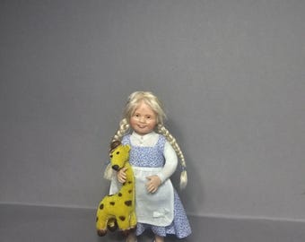 May include: A doll with blonde hair in two braids, wearing a blue and white floral print dress with a white apron. The doll is holding a small yellow plush giraffe toy with brown spots. The doll wears white shoes and has a smiling facial expression.