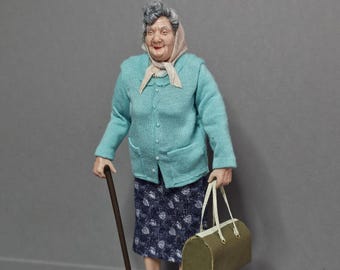 May include: A miniature figurine of an elderly person with gray hair wearing a light blue button-down cardigan, a dark blue patterned skirt, and a beige headscarf. The figure holds a brown walking cane and a green handbag, wearing blue shoes.