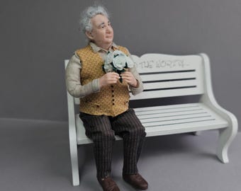 May include: A miniature figurine of an elderly person with gray hair, wearing a tan long-sleeve shirt, a yellow patterned vest, and dark pinstriped trousers, holding a small bouquet of white flowers while sitting on a white wooden bench.