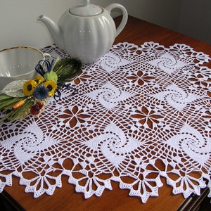 May include: White crocheted doily with a spiral pattern on a wooden table with a white teapot and a white cup and saucer.