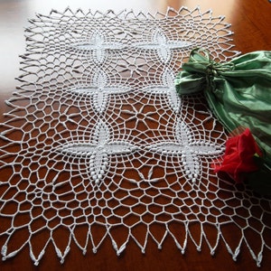 Può includere: Un runner da tavolo in crochet bianco con un delicato motivo di croci intrecciate. Il runner è posto su un tavolo di legno con una borsa di raso verde e una rosa rossa.