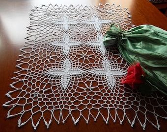 May include: White crocheted table runner with a delicate pattern of interwoven crosses. The runner is laid on a wooden table with a green satin bag and a red rose.
