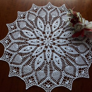 May include: White crocheted doily with a delicate floral pattern. The doily is round and has a scalloped edge.