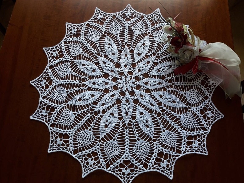 May include: White crocheted doily with a delicate floral pattern. The doily is round and has a scalloped edge.