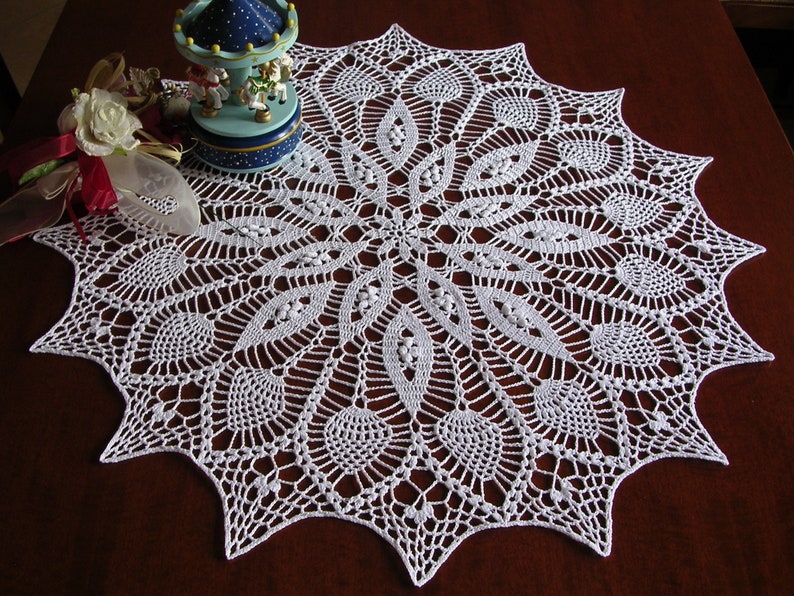 May include: White crocheted doily with a delicate, intricate design. The doily has a circular shape with a central motif and radiating patterns.