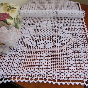 Puede incluir: Camino de mesa blanco de ganchillo con un delicado patrón floral. El camino de mesa está hecho con un hilo fino y tiene un borde festoneado.