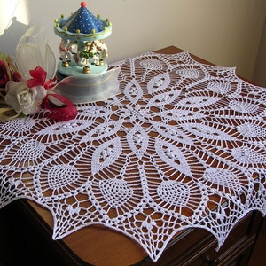 May include: White crocheted doily with a delicate, intricate design. The doily is laid on a wooden table with a small carousel figurine in the background.