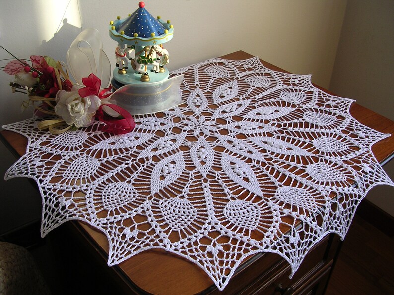 May include: White crocheted doily with a delicate, intricate design. The doily is laid on a wooden table with a small carousel figurine in the background.