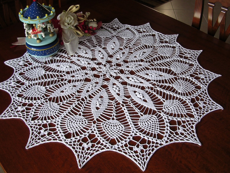 May include: White crocheted doily with a delicate, intricate design. The doily is circular with a scalloped edge and features a central motif of a starburst.