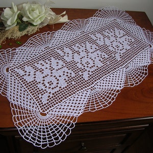 May include: A white, handmade crochet table runner with a detailed openwork design. The runner is rectangular with scalloped edges and a central pattern of geometric shapes. The runner is displayed on a dark wooden surface, with white flowers and greenery in the background.