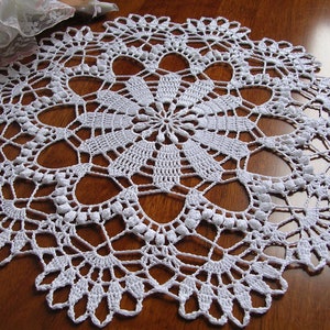 Puede incluir: Doily de ganchillo blanco con un delicado patrón floral. El doily es circular y tiene un borde festoneado.