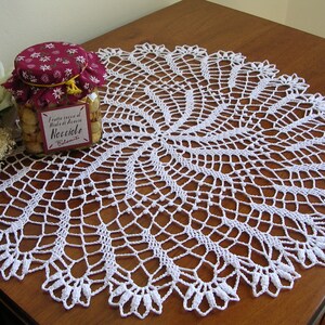 May include: White crocheted doily with a delicate, intricate design. The doily is laid on a wooden table with a jar of nuts in the background.