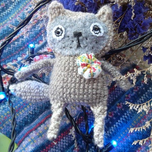 Può includere: Un peluche di gatto grigio lavorato a uncinetto con grandi occhi neri e un piccolo naso nero. Il gatto indossa un piccolo fiore bianco con un motivo floreale colorato. Il gatto è seduto su un tessuto blu e bianco a motivi con luci a stringa nere.