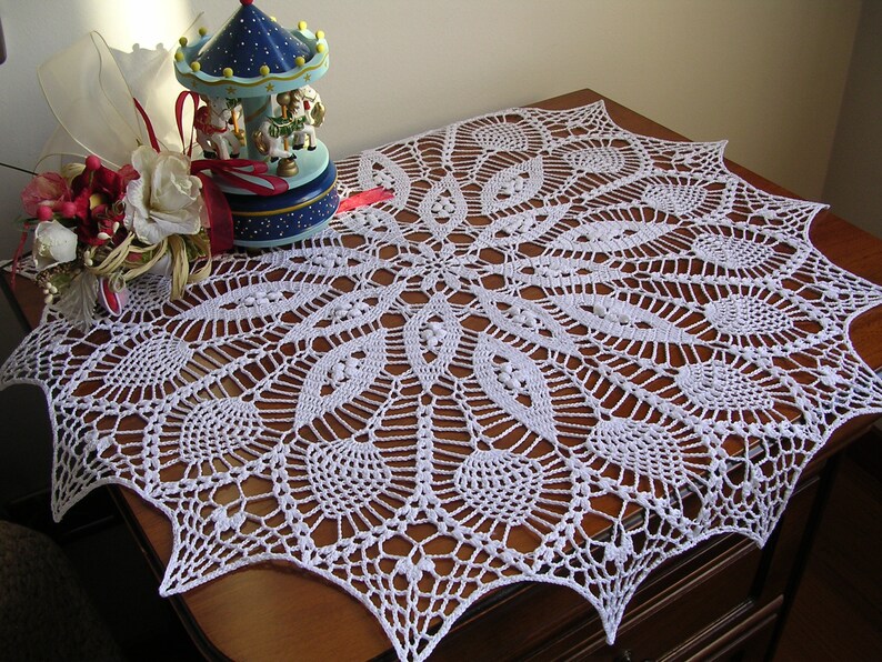May include: White crocheted table runner with a delicate, intricate design. The runner features a circular pattern with a starburst effect and a scalloped edge.