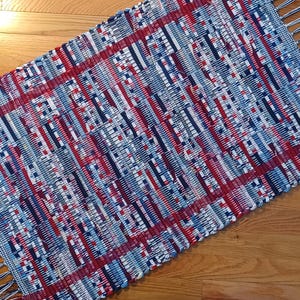 May include: Rectangular woven rug with a pattern of red, white, and blue stripes and blocks. The rug has a fringed edge and is made of a woven material. The rug is placed on a wooden floor.