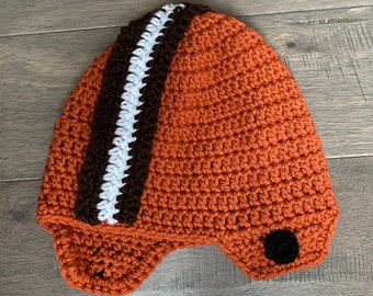 May include: A crocheted football helmet hat in orange, brown, and white. The hat has a black button on the side.