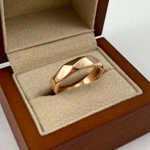 Puede incluir: Un anillo de banda de oro rosa con un dise&ntilde;o facetado y geom&eacute;trico. El anillo se muestra en una caja de madera.