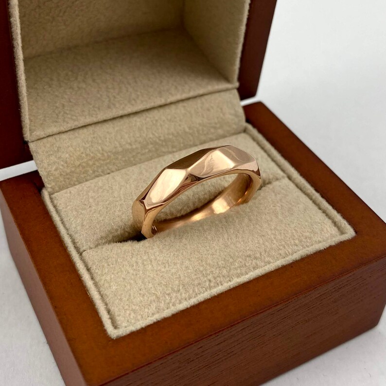 Puede incluir: Un anillo de banda de oro rosa con un dise&ntilde;o facetado y geom&eacute;trico. El anillo se muestra en una caja de madera.