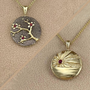 May include: Two gold pendants with red gemstone accents. One pendant features a stylized tree branch with three blossoms. The other pendant features a stylized sun with rays.