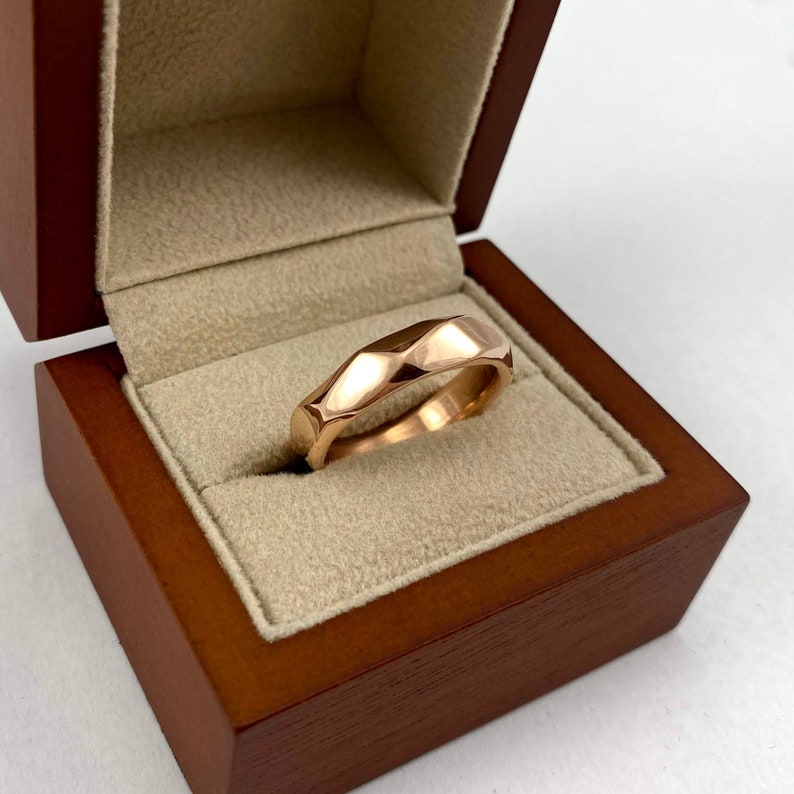 Puede incluir: Un anillo de banda de oro rosa con un dise&ntilde;o texturizado y facetado. El anillo se muestra en una caja de madera.