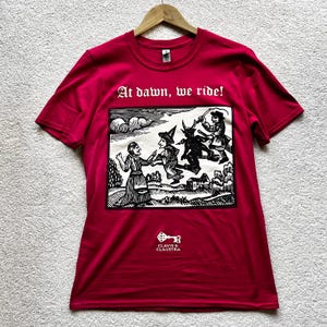 May include: A crimson red t-shirt with a black and white graphic print of witches riding brooms and the text "At dawn, we ride!". The shirt also features the text "CLAVIS & CLAUSTR".