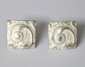 May include: Two square, silver-toned earrings with a textured, off-white surface. Each earring features a swirling, embossed design. The earrings are displayed against a plain, light background.