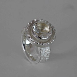 May include: A silver ring with a large, clear gemstone at the center. The gemstone is round and faceted, surrounded by a textured, circular setting. The band of the ring has a textured design. The ring is set against a light gray background.