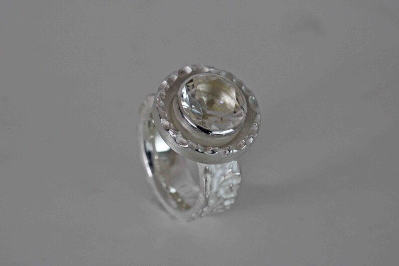 May include: A silver ring with a large, clear gemstone at the center. The gemstone is round and faceted, surrounded by a textured, circular setting. The band of the ring has a textured design. The ring is set against a light gray background.