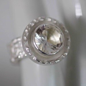 May include: A close-up of a silver ring with a large, clear, faceted gemstone. The ring has a textured band and a decorative bezel setting around the central stone. The ring is set against a soft, blurred background.