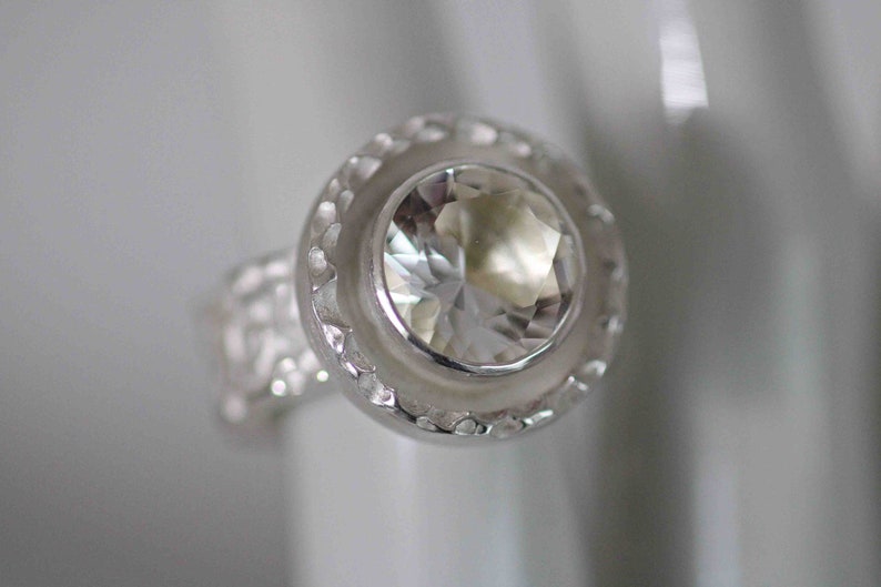 May include: A close-up of a silver ring with a large, clear, faceted gemstone. The ring has a textured band and a decorative bezel setting around the central stone. The ring is set against a soft, blurred background.