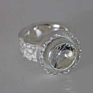 May include: A silver ring with a large, clear gemstone. The ring has a textured band and a scalloped bezel setting around the gemstone. The ring is displayed on a light gray surface.