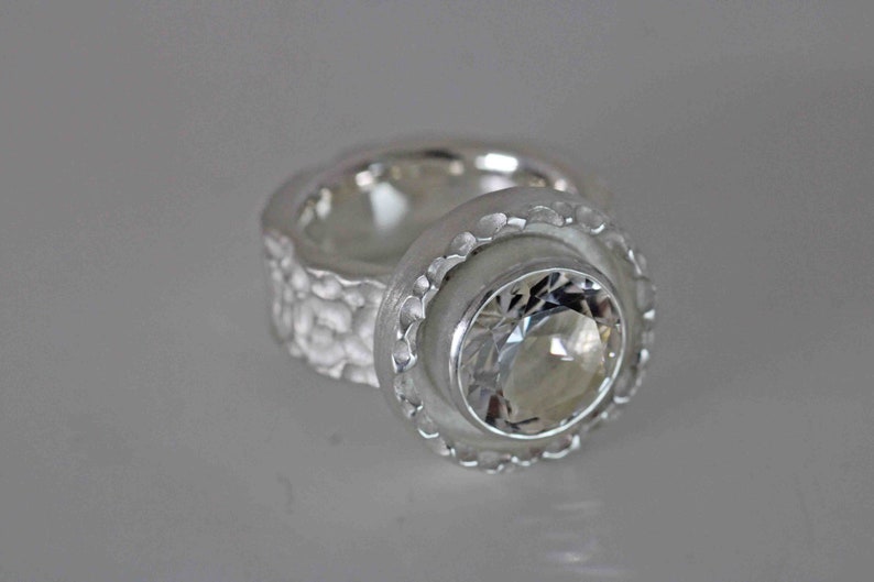 May include: A silver ring with a large, clear gemstone. The ring has a textured band and a scalloped bezel setting around the gemstone. The ring is displayed on a light gray surface.