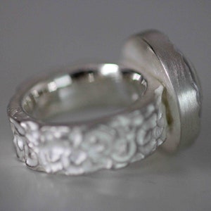 May include: A silver ring with a textured band and a large, rectangular setting. The band has a detailed, floral-like pattern. The setting has a brushed metal finish, and the ring is shown against a neutral background.
