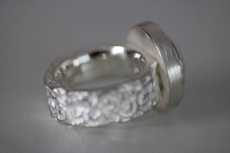 May include: A silver ring with a textured band and a large, rectangular setting. The band has a detailed, floral-like pattern. The setting has a brushed metal finish, and the ring is shown against a neutral background.