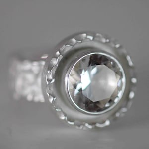 May include: A close-up of a silver ring with a large, clear, faceted gemstone. The ring has a textured band and a decorative bezel setting around the stone. The ring is set against a soft, gray background.