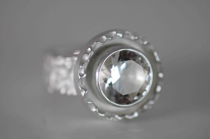May include: A close-up of a silver ring with a large, clear, faceted gemstone. The ring has a textured band and a decorative bezel setting around the stone. The ring is set against a soft, gray background.