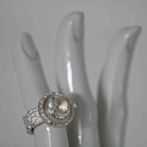 May include: A silver ring with a large, clear gemstone set in a halo design. The ring's band features a textured pattern. The ring is displayed on a white, hand-shaped display.