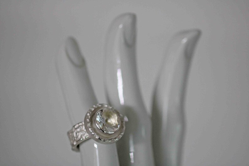 May include: A silver ring with a large, clear gemstone set in a halo design. The ring's band features a textured pattern. The ring is displayed on a white, hand-shaped display.