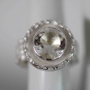 May include: A close-up of a silver ring with a large, clear, faceted gemstone. The ring has a textured, decorative band around the gemstone and a smooth, polished band. The ring is set against a white background.