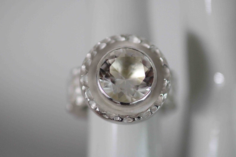 May include: A close-up of a silver ring with a large, clear, faceted gemstone. The ring has a textured, decorative band around the gemstone and a smooth, polished band. The ring is set against a white background.